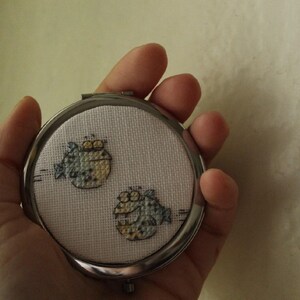 May include: A silver compact mirror with a white background and two embroidered blue and yellow fish.