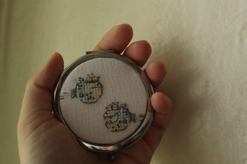 May include: A silver compact mirror with a white background and two embroidered blue and yellow fish.