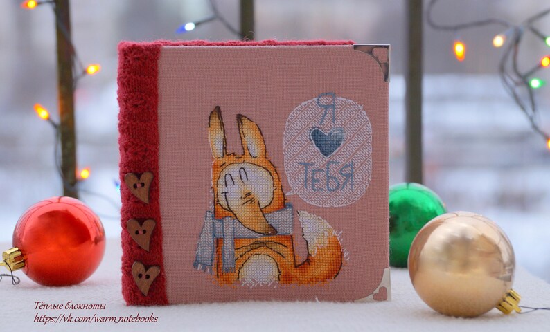 Puede incluir: Un cuaderno rosa con un borde de punto rojo y botones de madera en forma de coraz&oacute;n. La portada presenta un zorro bordado con una bufanda azul y un globo de di&aacute;logo que dice "Te amo" en ruso.