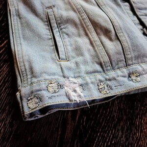 May include: Light blue denim jacket with a distressed hem and embroidered details. The jacket has a single pocket on the left side.
