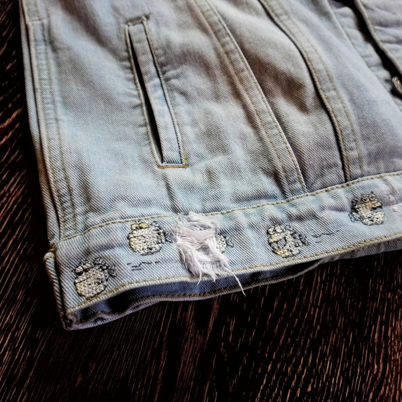 May include: Light blue denim jacket with a distressed hem and embroidered details. The jacket has a single pocket on the left side.