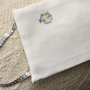 May include: White fabric with a small embroidered image of a yellow fish with blue fins. The fabric is attached to a silver sequined trim.