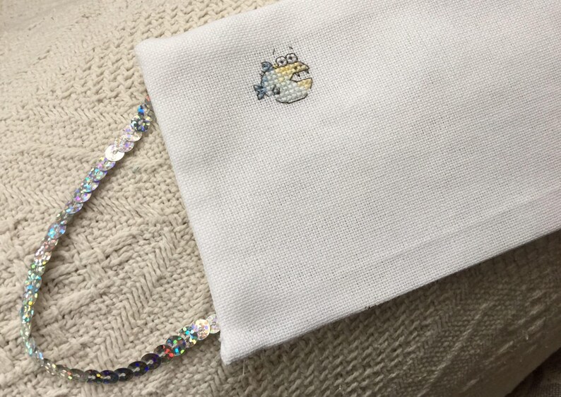 May include: White fabric with a small embroidered image of a yellow fish with blue fins. The fabric is attached to a silver sequined trim.