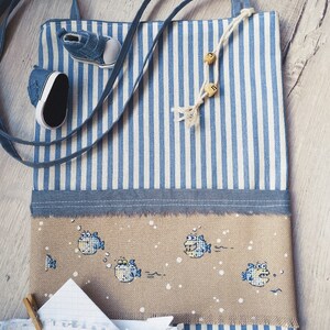 May include: A blue and white striped tote bag with a cross-stitch design of fish on the front. The bag has a blue strap and a paper note with the text "#stayhome" attached to it.