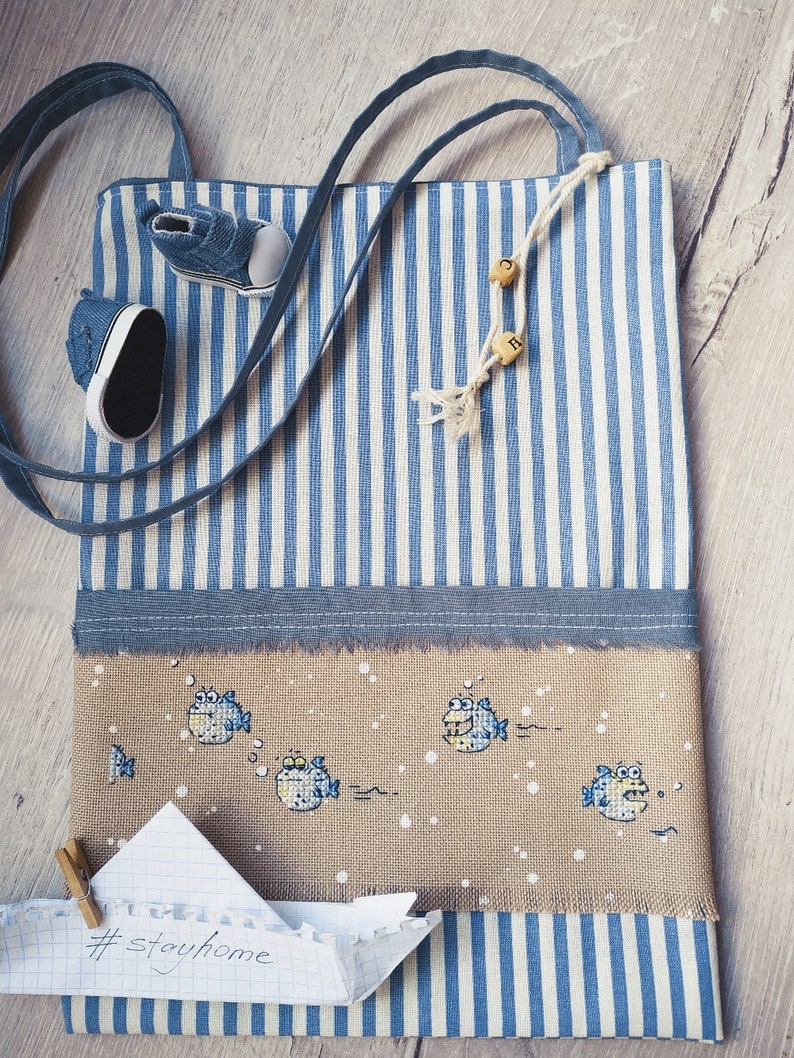 May include: A blue and white striped tote bag with a cross-stitch design of fish on the front. The bag has a blue strap and a paper note with the text "#stayhome" attached to it.