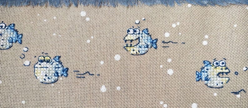 May include: A beige fabric with four blue and yellow fish embroidered on it. The fish have white eyes and are swimming in different directions. There are white dots scattered across the fabric.