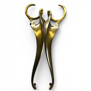 May include: A pair of decorative brass scissors. The scissors have a distinctive design, with curved handles and blades. The brass has a polished, golden finish. The scissors are set against a white background. The overall design is elegant and artistic.