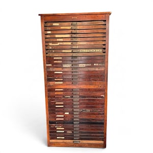 May include: A tall, dark brown wooden cabinet featuring numerous horizontal drawers. Each drawer has a small metal pull handle and a label holder. Some drawers display printed text labels, including ANGLO-FRENCH CAP and JUMBO.