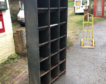 May include: A vintage metal industrial shelving unit with multiple compartments. The shelves are painted a dark green color and have a rustic look. The unit is standing on a paved surface.