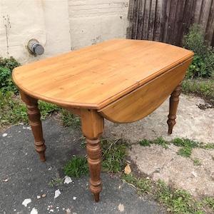 May include: A light brown, antique drop-leaf table with a rounded edge. The table has four turned legs and is made of wood. The table is shown with one leaf extended, and the other folded down. The table is approximately 122 cm long.