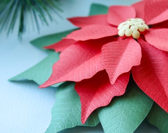 Könnte beinhalten: Eine rote Papier-Weihnachtsstern-Blume mit grünen Papierblättern. Die Blume besteht aus mehreren Papierschichten, die einen 3D-Effekt erzeugen.