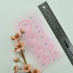 Op de afbeelding: Een transparante plastic plaat met 10 ronde, roze, verhoogde cirkels in verschillende maten. De cirkels zijn gelabeld met metingen in millimeters. Een liniaal is zichtbaar op de achtergrond.