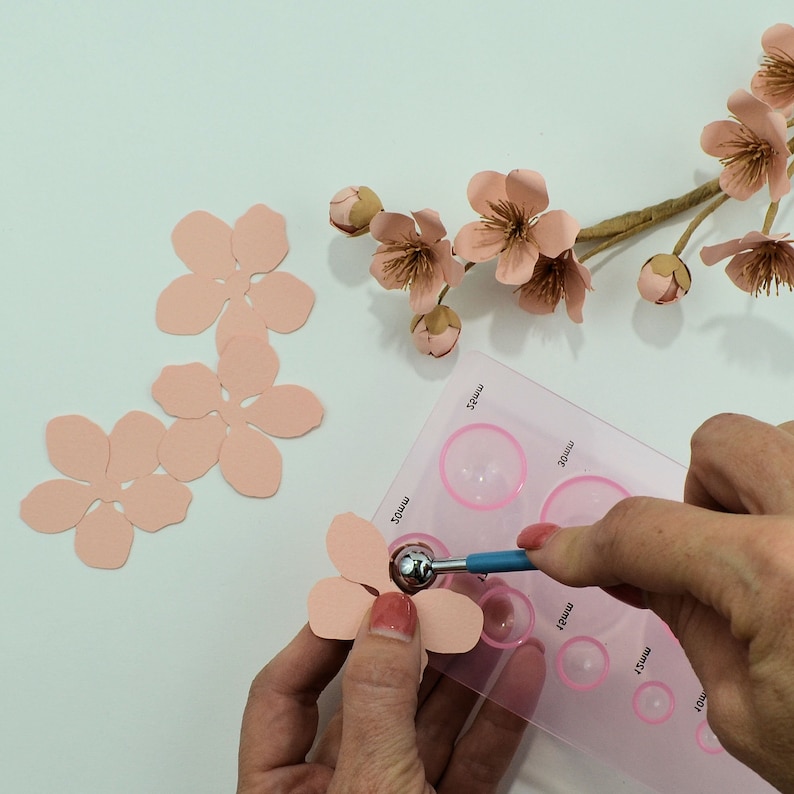 Op de afbeelding: Een close-up van een persoon die een gereedschap gebruikt om een ​​deuk in een roze papieren bloemblaadje te maken. Het gereedschap wordt gebruikt op een roze plastic mat met cirkels van verschillende groottes die in millimeters zijn gelabeld.