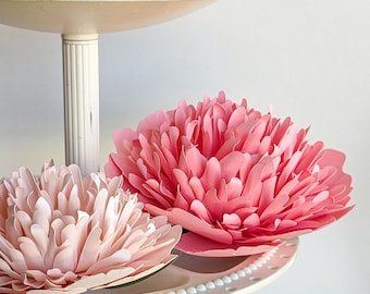 May include: Two pink paper flowers on a white tiered cake stand. The flowers are made of paper and have a delicate, ruffled texture.