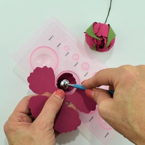 Op de afbeelding: Een close-up van iemand die een gereedschap gebruikt om een ​​bloemblaadje van bordeauxrood papier te vormen. Het bloemblaadje wordt gevormd over een ronde mal die is gemarkeerd met metingen in millimeters.