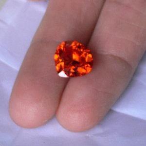 Może przedstawiać: Jasny pomarańczowy kamień szlachetny o fasetowanym szlifie. Kamień szlachetny jest trzymany między dwoma palcami.