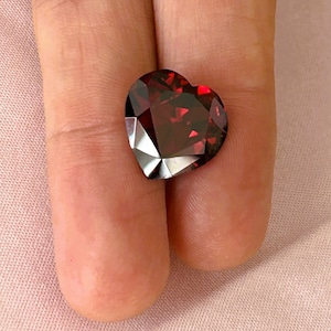 Może przedstawiać: Fasetowany, w kształcie serca kamień szlachetny w głębokim odcieniu czerwieni. Kamień ma brylantowy szlif, odbijający światło wieloma fasetami. Kształt serca jest wyraźnie zdefiniowany, z ostrymi kątami i wypolerowaną powierzchnią. Kamień szlachetny ma bogaty, ciemnoczerwony kolor.