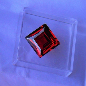 Może przedstawiać: Czerwony kamień szlachetny o kwadratowym szlifie, prezentowany w przezroczystej plastikowej pudełku.