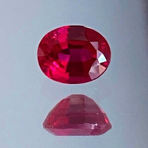 May include: A vibrant red oval-shaped gemstone with a faceted cut. The gemstone is displayed on a white background.