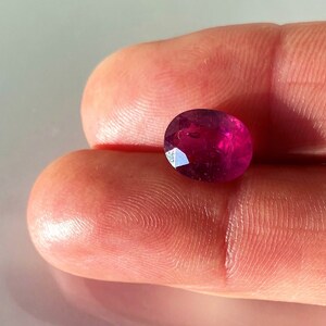 May include: A vibrant, oval-shaped ruby gemstone with a deep pink hue. The gemstone is set against a neutral background.