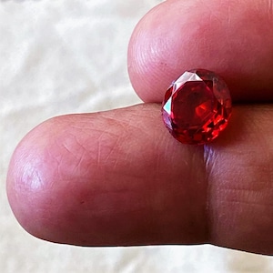 Może przedstawiać: Fasetowany, owalny, czerwony kamień szlachetny. Klejnot trzymany jest między dwoma palcami, prezentując swój brylantowy szlif i żywy kolor. Fasetki kamienia szlachetnego odbijają światło, tworząc efekt iskrzenia. Klejnot ma głęboki, bogaty czerwony kolor.
