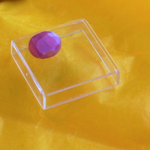 May include: A clear plastic square display case with a single, large, faceted, purple gemstone inside.