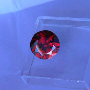 Może przedstawiać: Okrągły, fasetowany, głęboko czerwony kamień szlachetny, prawdopodobnie granat lub rubin, jest prezentowany w przezroczystym, kwadratowym plastikowym pudełku. Kamień ma wiele faset, które odbijają światło, tworząc efekt iskrzenia.