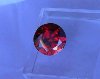 May include: A round, faceted, deep red gemstone, possibly a garnet or ruby, is displayed in a clear, square plastic case. The gem has multiple facets that reflect light, creating a sparkling effect. The background is a gradient of blue.