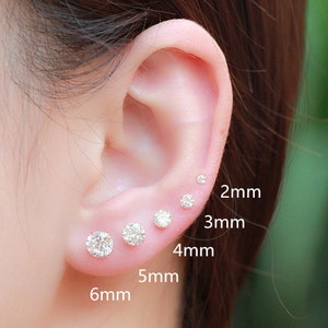 May include: A close-up of an ear with six different sized clear round stud earrings. The earrings are labeled with their size in millimeters: 2mm, 3mm, 4mm, 5mm, and 6mm.