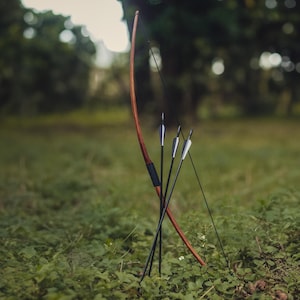 May include: A wooden bow and arrows are placed in a grassy field. The bow is brown and the arrows are black with silver tips.