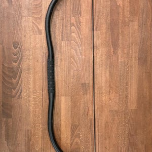 May include: A black recurve bow with a textured grip. The bow is made of a dark material and has a black string. The bow is set against a wooden background.