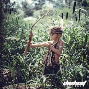 May include: A young person dressed in a Native American inspired costume holds a bow and arrow in a grassy field. The person is wearing a beaded necklace and a headdress with feathers. The bow is made of wood and the arrow is tipped with a brown point.