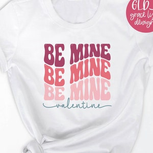 May include: White t-shirt with the text "BE MINE" repeated three times in a retro font, with the words stacked on top of each other. The text is in shades of pink, red, and purple. The words "valentine" are written in a smaller font below the stacked text.