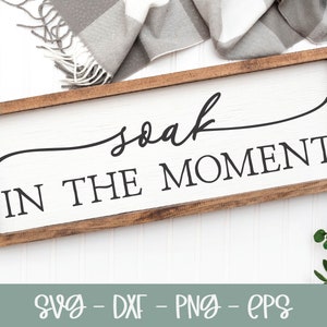 May include: A wooden sign with a white background and brown frame. The sign says "soak in the moment" in black cursive lettering.