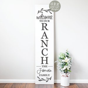 Puede incluir: Un letrero de madera blanco con letras negras que dice "Bienvenido a nuestro rancho - La familia Johnson". El letrero está decorado con una corona de flores en la parte superior e inferior.