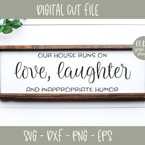 May include: A brown wooden sign with white text that reads "Our house runs on love, laughter and inappropriate humor". The sign is on a white wooden background with a black and white checkered ribbon and green foliage.