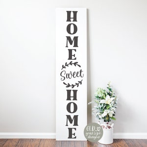 May include: A tall, white wooden sign with the words "HOME SWEET HOME" in black lettering. The word "Sweet" is within a circular wreath design. A small bouquet of white flowers sits in a white bucket to the right.