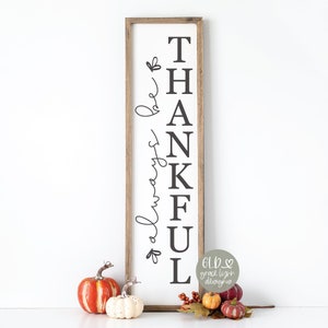 Può includere: Un cartello di legno con le parole "Be Always Thankful" scritte in lettere nere. Il cartello è appoggiato a un muro bianco e davanti a esso ci sono piccole zucche e foglie autunnali.