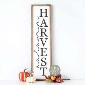 May include: A wooden sign with the word "HARVEST" written vertically in black letters. The word "autumn" is written in cursive script to the left of the letters. The sign is sitting on a white surface with a few small pumpkins and fall leaves in front of it.