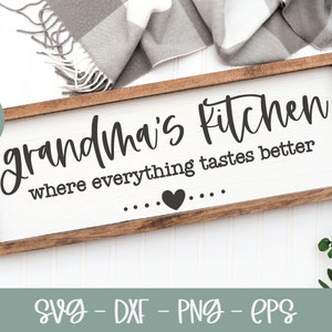 May include: A wooden sign with the text "Grandma's Kitchen where everything tastes better" in black script font. A small heart is centered below the text. The sign is on a white surface with a gray blanket and green foliage.