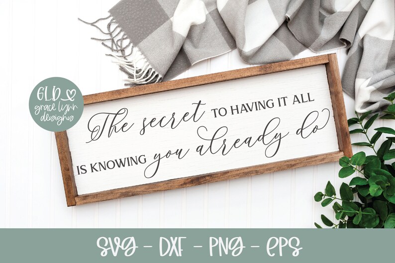 May include: A rectangular wooden sign with the quote "The secret to having it all is knowing you already do" in elegant script. The sign has a white background and a dark brown frame. The image also includes a gray and white plaid blanket and greenery.