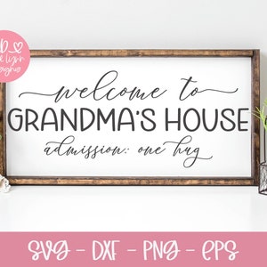 May include: A white sign with a brown wooden frame, featuring the text "Welcome to Grandma's House, admission: one hug". The sign is displayed on a white surface with a pink flower and a potted plant in the background.