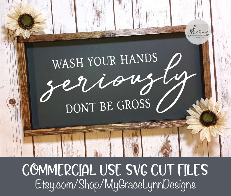 May include: A dark gray wooden sign with white text that reads "Wash your hands seriously don't be gross". The sign is framed in a dark brown wood frame and is surrounded by white wood paneling.