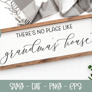May include: A wooden sign with the text "THERE'S NO PLACE LIKE GRANDMA'S HOUSE" in black cursive font. The sign is on a white surface with a gray and white blanket and green leaves in the background.