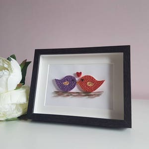 May include: A framed paper craft artwork featuring two birds, one purple and one red, with a small red heart above them. The birds are kissing and the artwork is set against a white background.