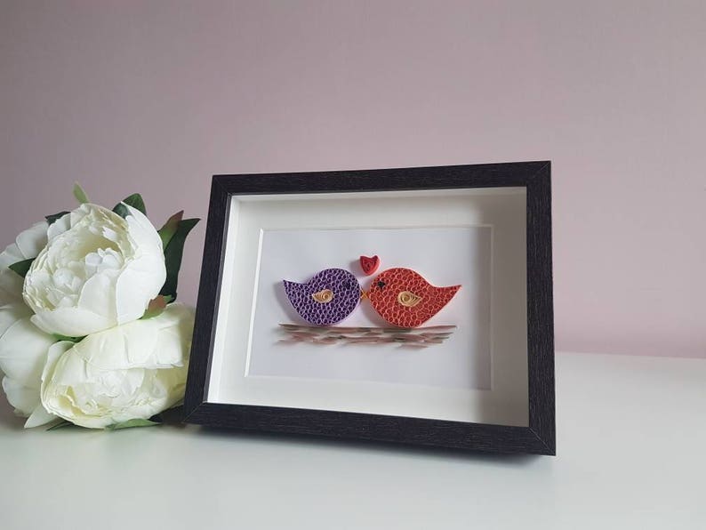 May include: A framed paper craft artwork featuring two birds, one purple and one red, with a small red heart above them. The birds are kissing and the artwork is set against a white background.