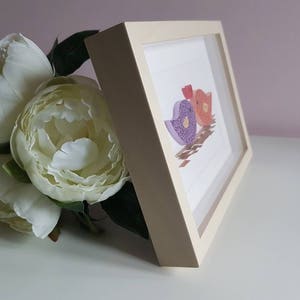 May include: A white frame with a light pink, purple, and orange paper bird design inside. The frame is sitting on a white surface.