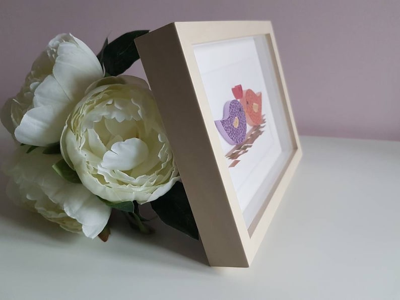 May include: A white frame with a light pink, purple, and orange paper bird design inside. The frame is sitting on a white surface.