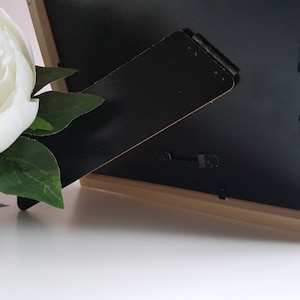 May include: A black picture frame with a wooden frame and a black easel stand. The frame is sitting on a white surface.