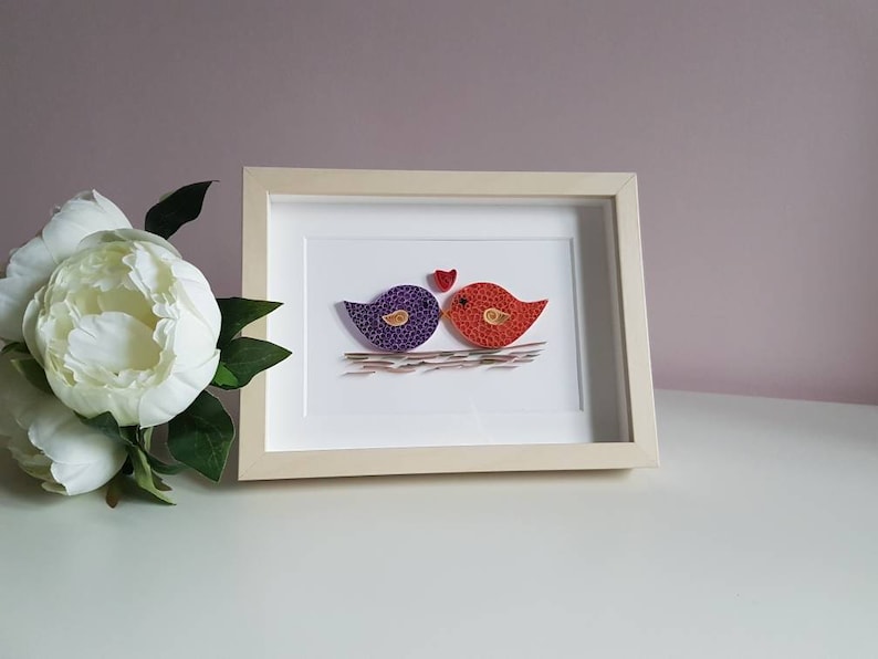 May include: A framed paper craft artwork featuring two birds, one purple and one orange, kissing with a red heart above them. The birds are made of paper and are in a simple nest.